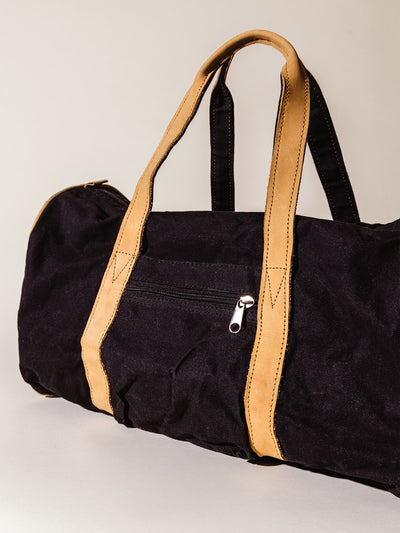 Back view of a black canvas duffel bag featuring a zippered pocket and tan leather handles, photographed in soft studio lighting.