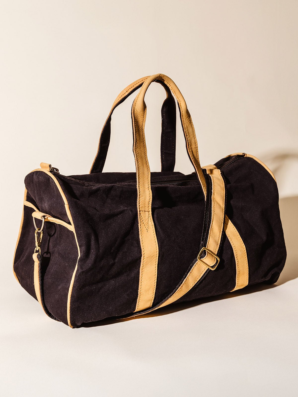 Angled view of a black and tan canvas duffel bag with leather trim and detachable shoulder strap, set against a beige backdrop.