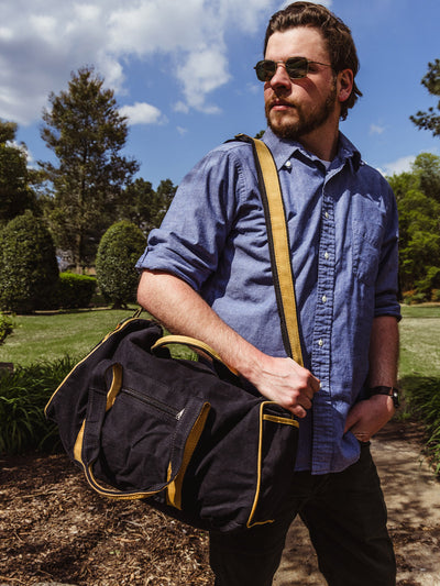 A man wearing sunglasses, a blue button-up shirt, and dark pants stands confidently outdoors with a black canvas and tan leather-trimmed duffel bag slung over his shoulder. Trees and a partly cloudy sky fill the background.