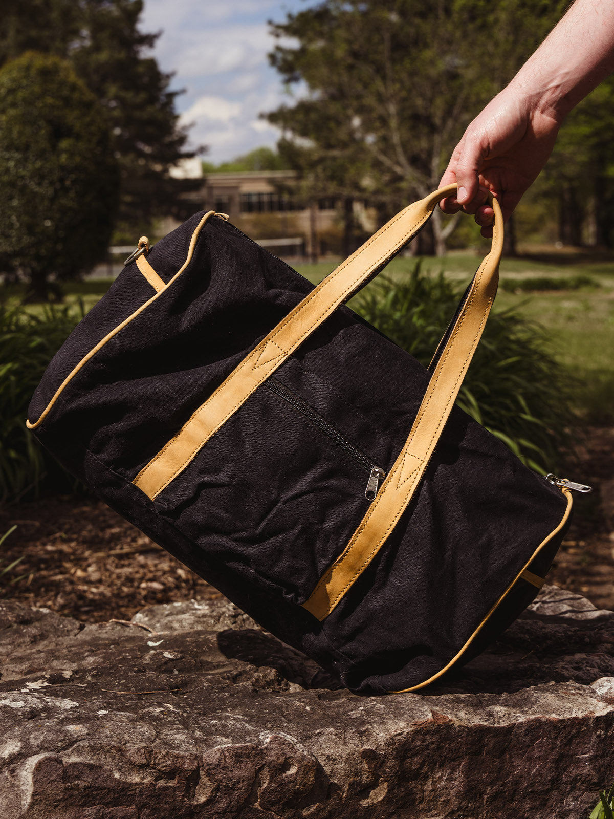 Close-up of a hand holding a black canvas duffel bag with tan leather handles, resting on a large rock in a sunny park setting.