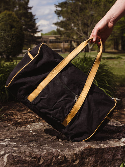 Close-up of a hand holding a black canvas duffel bag with tan leather handles, resting on a large rock in a sunny park setting.