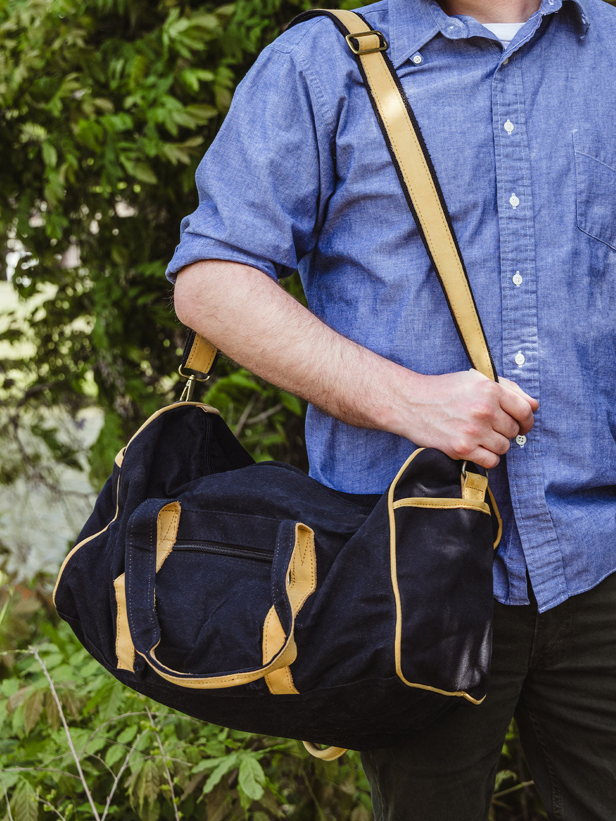 A man in a blue shirt holds a black and tan duffel bag at his side using the adjustable shoulder strap. He stands in front of lush greenery.