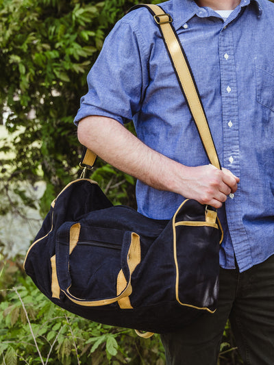 A man in a blue shirt holds a black and tan duffel bag at his side using the adjustable shoulder strap. He stands in front of lush greenery.