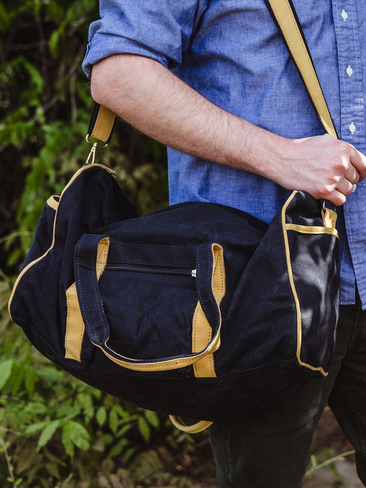 The same duffel bag is shown from a slightly different angle, slung over the man's shoulder with the zippered pocket and stitching details clearly visible.