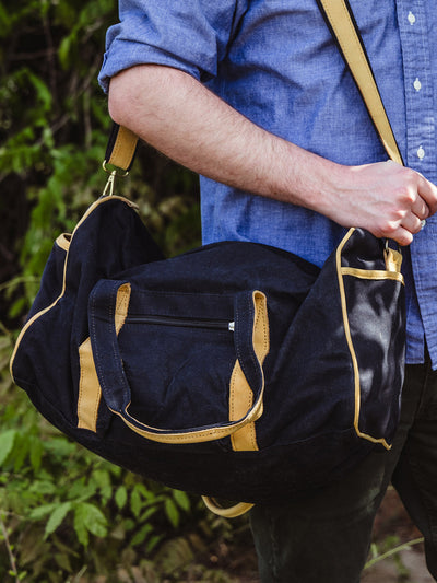 The same duffel bag is shown from a slightly different angle, slung over the man's shoulder with the zippered pocket and stitching details clearly visible.