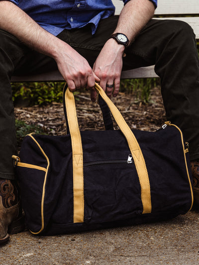 A man sits on a bench with a duffel bag resting in front of him on the ground. He holds the tan handles with both hands. He wears a blue shirt, dark green pants, a wristwatch, and cowboy boots.