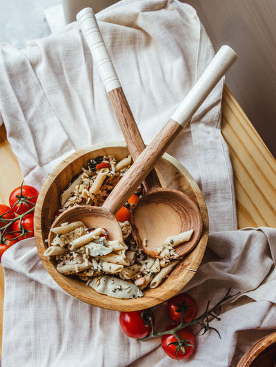 Handcrafted wooden salad servers with white-wrapped handles resting in a natural wooden bowl filled with pasta and vegetables, surrounded by vine tomatoes on a soft linen cloth, emphasizing the rustic charm and functional beauty of artisanal kitchen essentials.