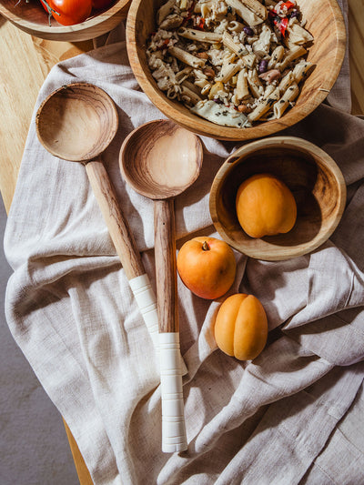 Two handcrafted wooden salad servers with white-wrapped handles resting on a natural linen cloth, next to a rustic wooden bowl filled with pasta salad and a smaller bowl holding fresh apricots, capturing the warmth and artisan craftsmanship of handmade kitchenware.