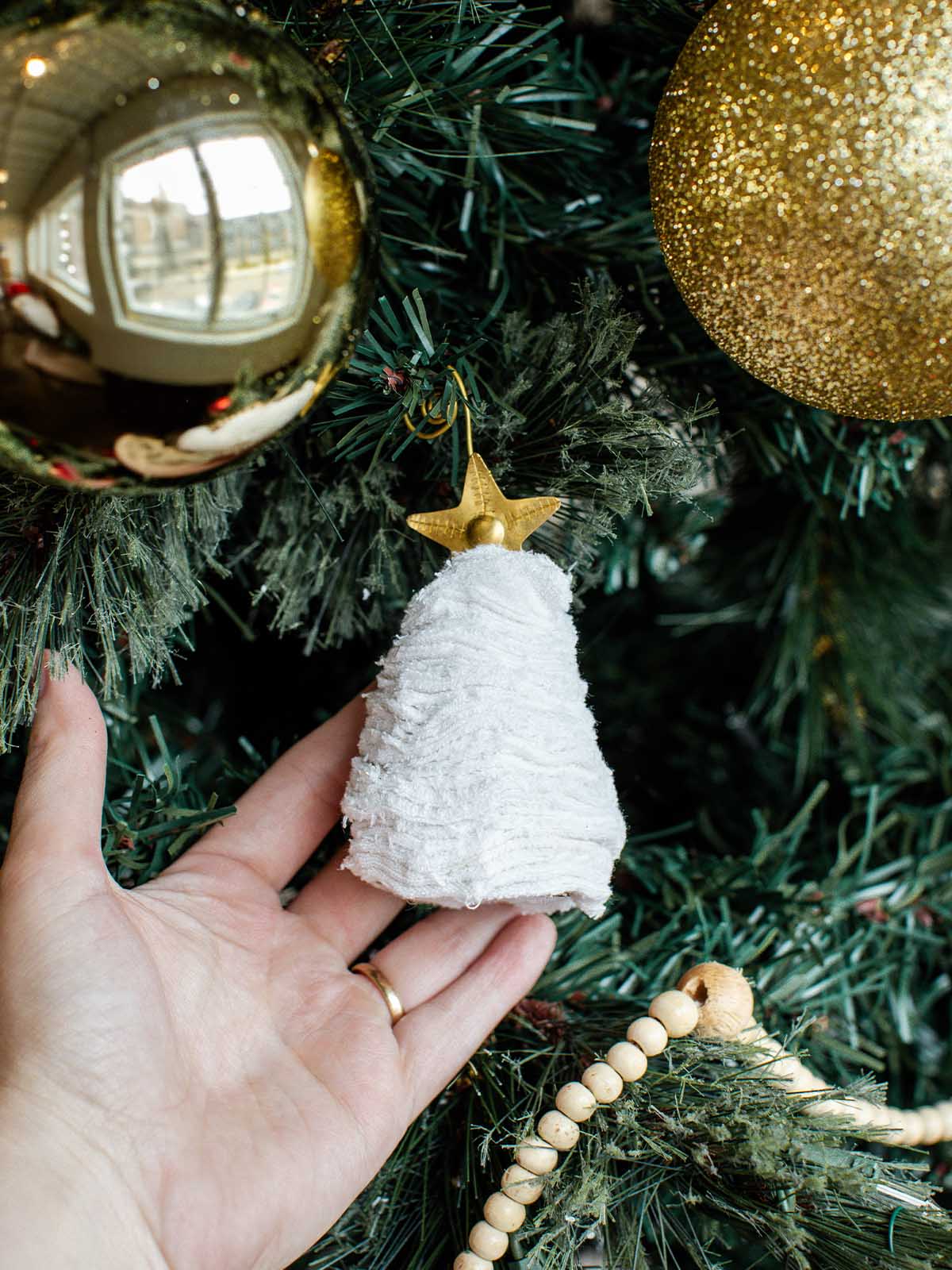 Hand holding a handmade white Christmas tree ornament with a gold star topper, hung on a decorated Christmas tree with gold baubles.