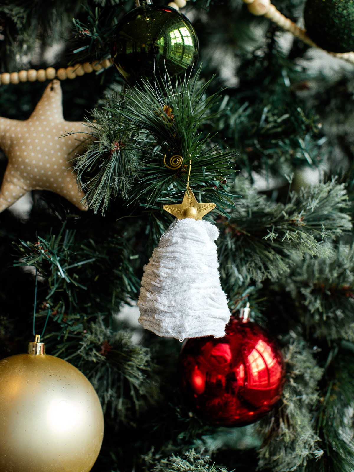 White fabric Christmas tree ornament with gold star hanging among red, gold, and green baubles on a festive holiday tree.