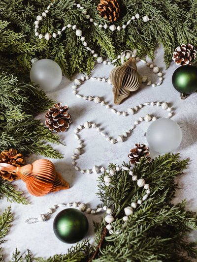 Flat lay of handmade white paper bead Christmas garland styled with pine greenery, pinecones, frosted glass ornaments, and metallic paper ornaments — ethical holiday décor.