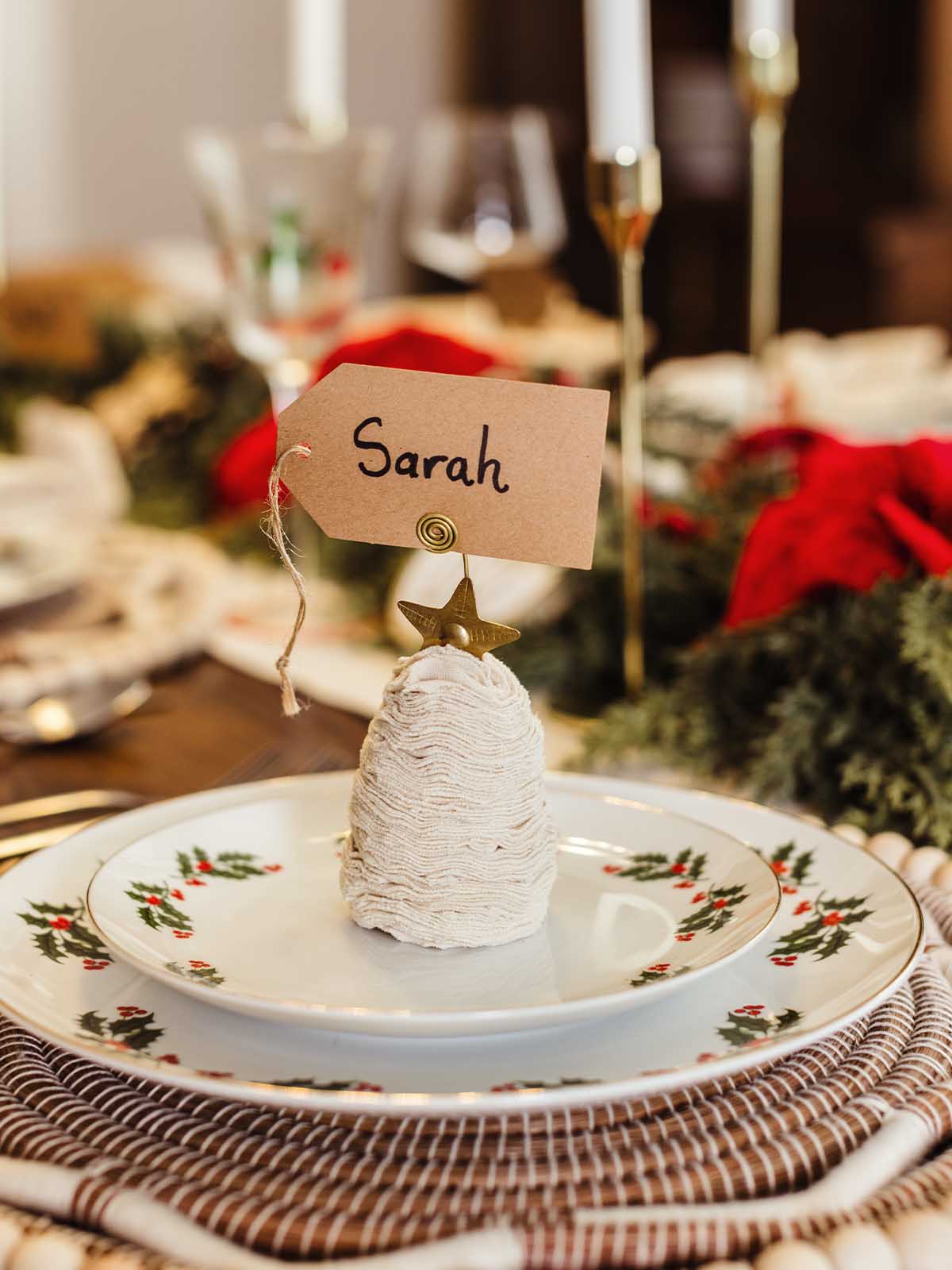 Handcrafted sweater Christmas tree ornament used as a festive place card holder with a star topper and name tag reading ‘Sarah,’ styled on a holiday table setting with holly-patterned china and woven placemat.