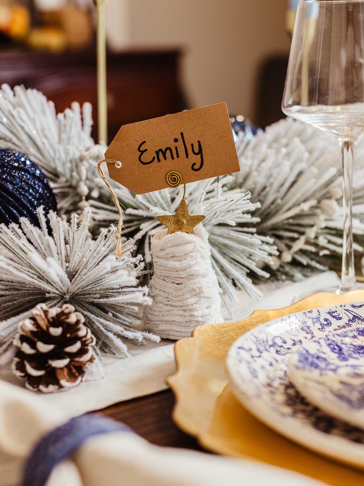 Cozy sweater Christmas tree ornament with gold star topper holding a name tag reading ‘Emily,’ placed on an elegant winter tablescape with frosted greenery, pinecones, and layered gold and blue dinnerware.