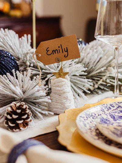 Cozy sweater Christmas tree ornament with gold star topper holding a name tag reading ‘Emily,’ placed on an elegant winter tablescape with frosted greenery, pinecones, and layered gold and blue dinnerware.