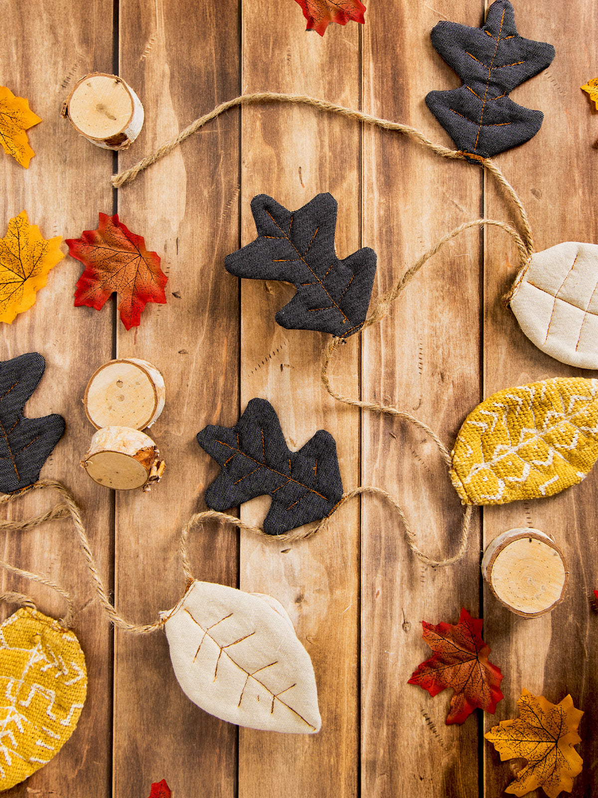 Handmade fabric leaf garland in black, cream, and yellow patterns on a jute string, displayed on a rustic wooden surface with scattered decorative autumn leaves and wood slices.
