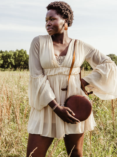 Female African model in cream dress holding the canteen purse with her hand while the purse is over her shoulder. Model and purse are in an outdoor setting. 