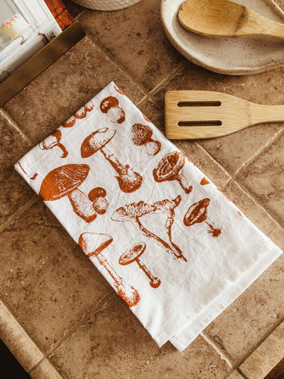 White cotton tea towel with hand-drawn rust-colored mushroom illustrations, neatly folded on a tiled kitchen counter. Wooden cooking utensils and a ceramic plate rest nearby.
