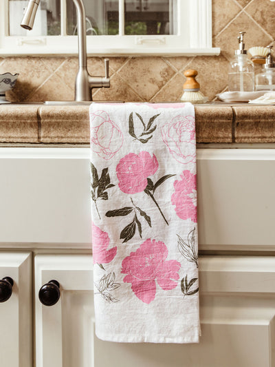 White cotton tea towel printed with large pink peonies and green leaves, hanging over a rustic stone kitchen sink. A modern stainless steel faucet and soap dispensers are visible in the background.