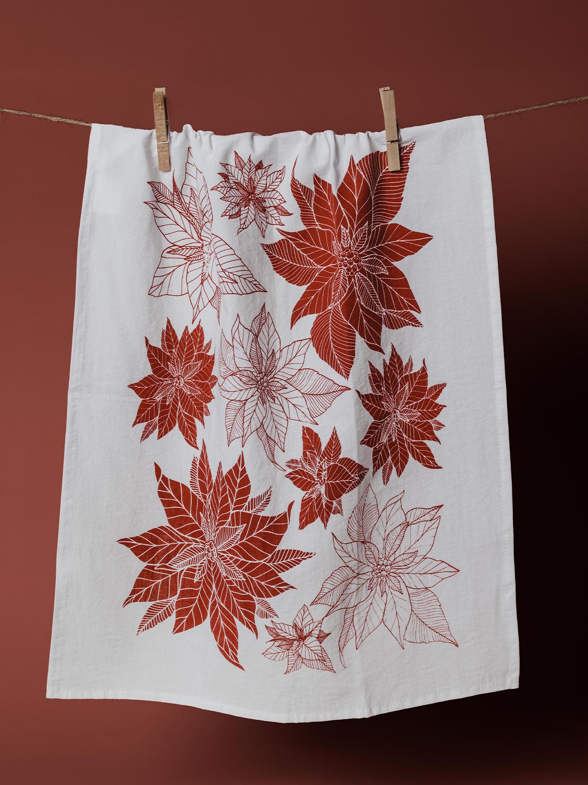 White kitchen towel featuring a red poinsettia print, hanging on a clothesline with a green background, perfect for holiday decor and festive kitchens.