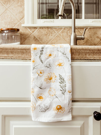 White cotton tea towel featuring a delicate yellow and green floral pattern draped over a farmhouse sink with a stone countertop and brushed nickel faucet. A glass jar sits in the background near a window.