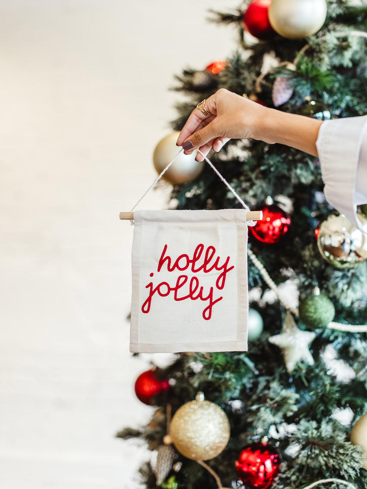 Hand holding a small hanging canvas banner with red “holly jolly” text in front of a decorated Christmas tree with ornaments.