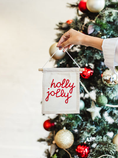 Hand holding a small hanging canvas banner with red “holly jolly” text in front of a decorated Christmas tree with ornaments.