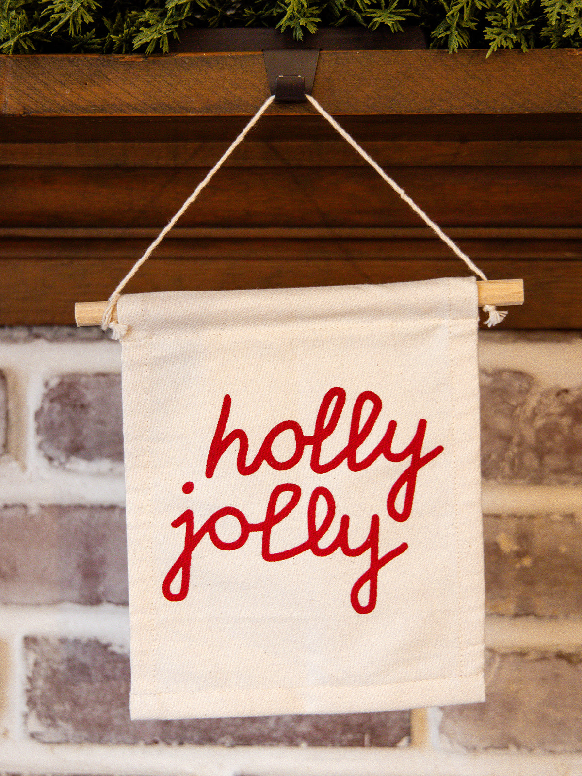Close-up of holiday “holly jolly” canvas wall hanging with red lettering, styled on a rustic brick fireplace mantel with greenery.