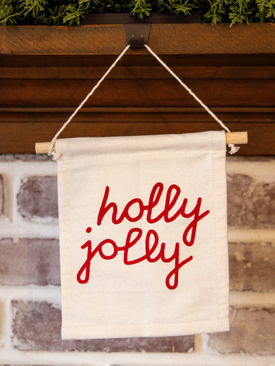 Close-up of holiday “holly jolly” canvas wall hanging with red lettering, styled on a rustic brick fireplace mantel with greenery.