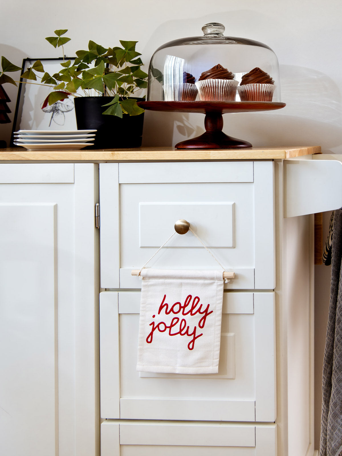 Festive “holly jolly” hanging banner displayed on a white kitchen cabinet, styled with cupcakes under a glass dome and potted plant decor.