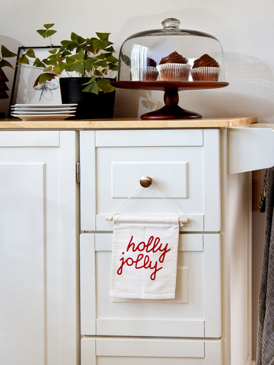 Festive “holly jolly” hanging banner displayed on a white kitchen cabinet, styled with cupcakes under a glass dome and potted plant decor.