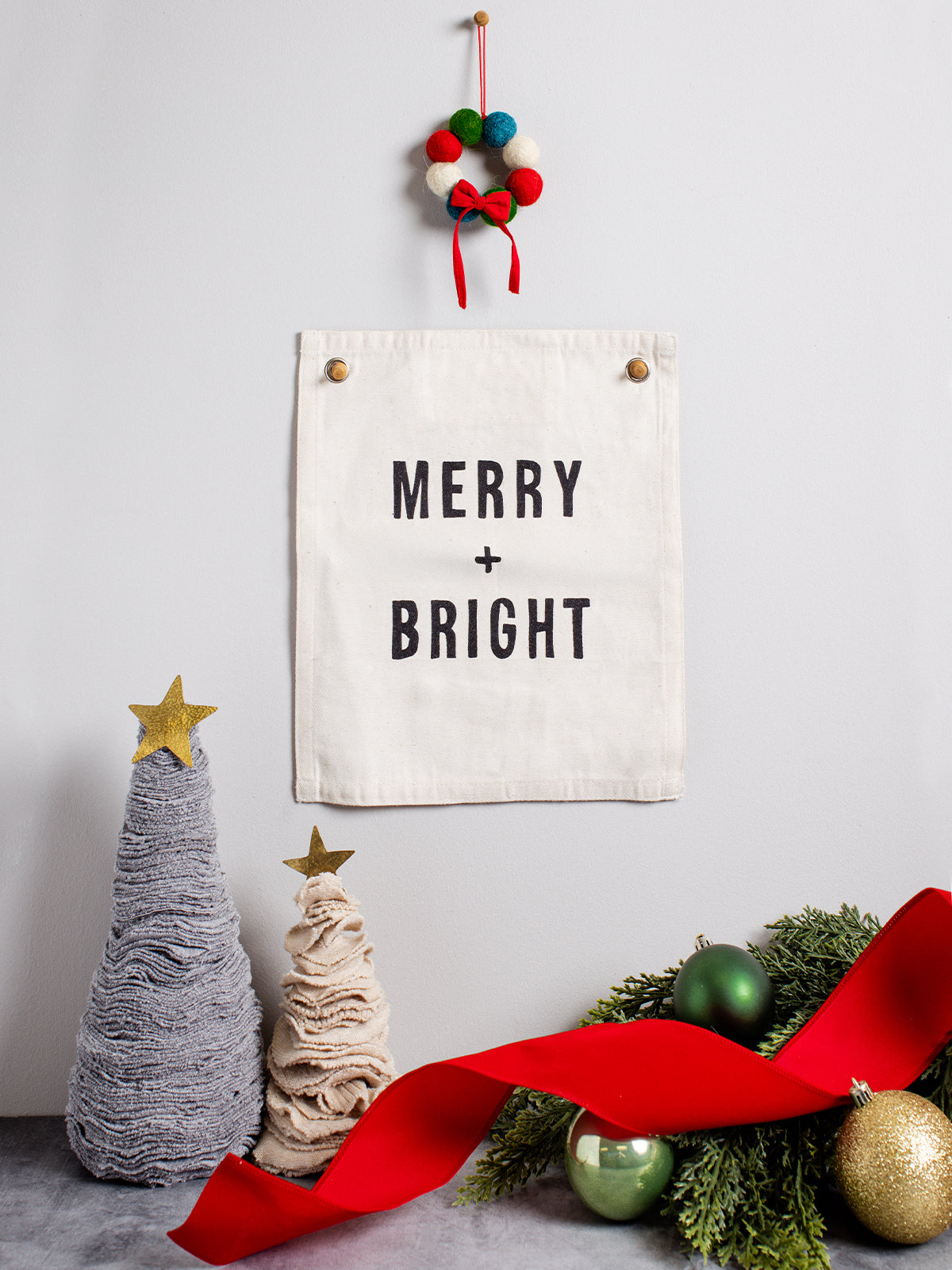 Minimal holiday wall décor with a cotton banner reading ‘MERRY + BRIGHT’ hung on a light gray wall, styled with felt ball wreath, neutral fabric Christmas trees topped with gold stars, evergreen sprigs, green and gold ornaments, and a flowing red ribbon.