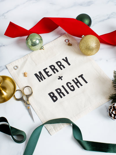 Flat lay of a neutral cotton ‘MERRY + BRIGHT’ holiday banner surrounded by festive wrapping accents — red and green velvet ribbon, gold ornaments, evergreen sprigs, pinecones, scissors, and a brass candle holder — perfect for Christmas decorating or gifting.