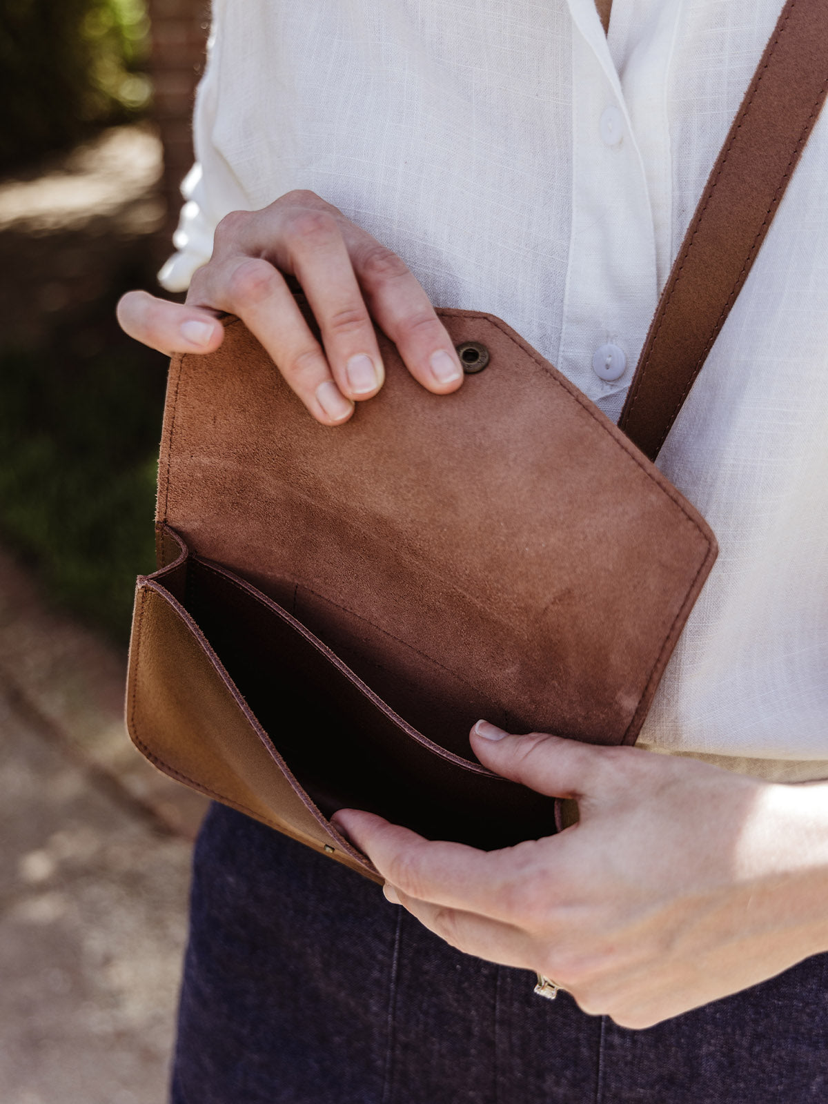 Close-up of a woman opening a brown leather crossbody bag with a suede flap and snap closure, wearing a white blouse.