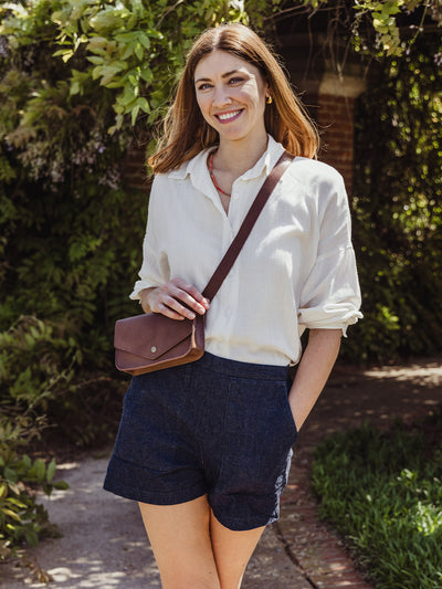 Woman standing outdoors smiling, wearing a white blouse, navy shorts, and a brown leather crossbody bag from JOYN.