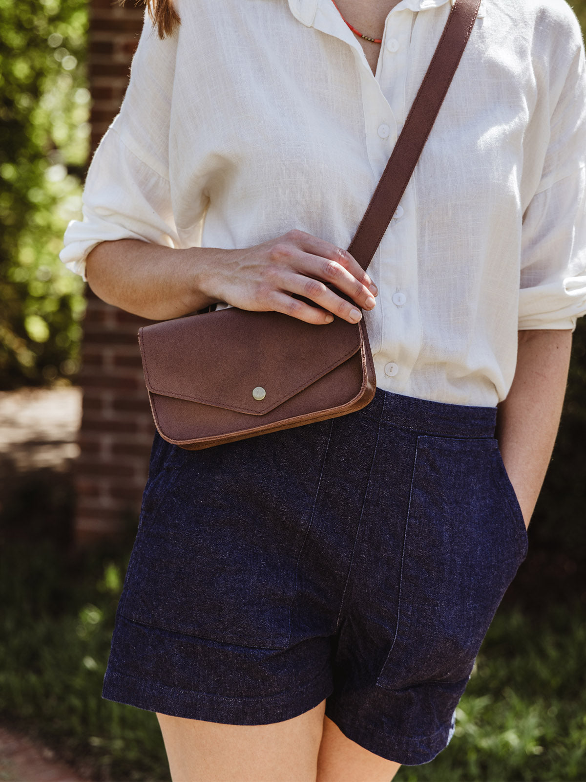 Cropped view of a woman wearing a white button-up blouse and navy shorts, showcasing a brown leather crossbody bag worn over the shoulder.
