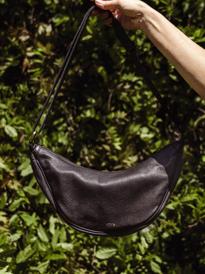 Hand holding a black leather crescent crossbody bag with a pebbled texture and adjustable strap against a leafy background.