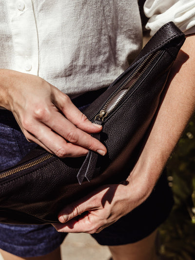 Close-up of a woman zipping the black leather crescent bag, showcasing the bag’s durable zipper and textured finish.