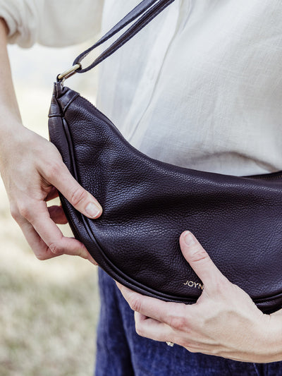 Woman holding a black leather crescent crossbody bag with visible JOYN logo, worn over a white blouse and navy shorts.