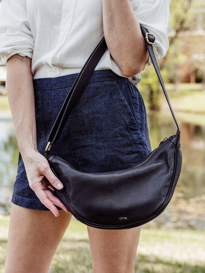 Cropped view of a woman modeling the black leather crescent crossbody bag by JOYN, with gold hardware and structured shape.
