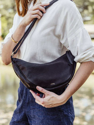 Close-up of a woman wearing a white blouse and holding a black leather crescent crossbody bag by JOYN with gold logo detail, styled with dark blue high-waisted shorts.