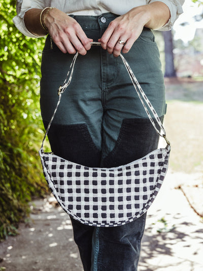 Close-up of a woman holding a black-and-white checkered crossbody bag by its strap in front of her green pants.