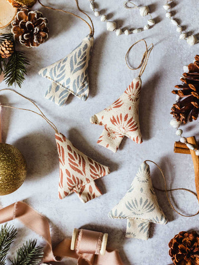Set of four handmade fabric Christmas tree ornaments with red and blue block print designs, styled with pinecones, gold bauble, ribbon, and festive greenery.