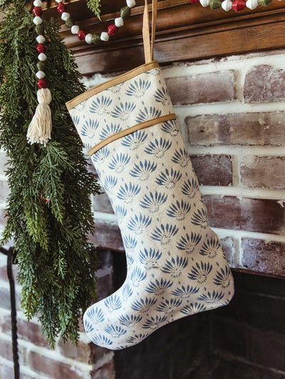 Single blue hand-block printed Christmas stocking with botanical flower pattern hanging from a rustic brick fireplace mantel, accented with evergreen garland and wood bead holiday garland — sustainable Christmas styling.