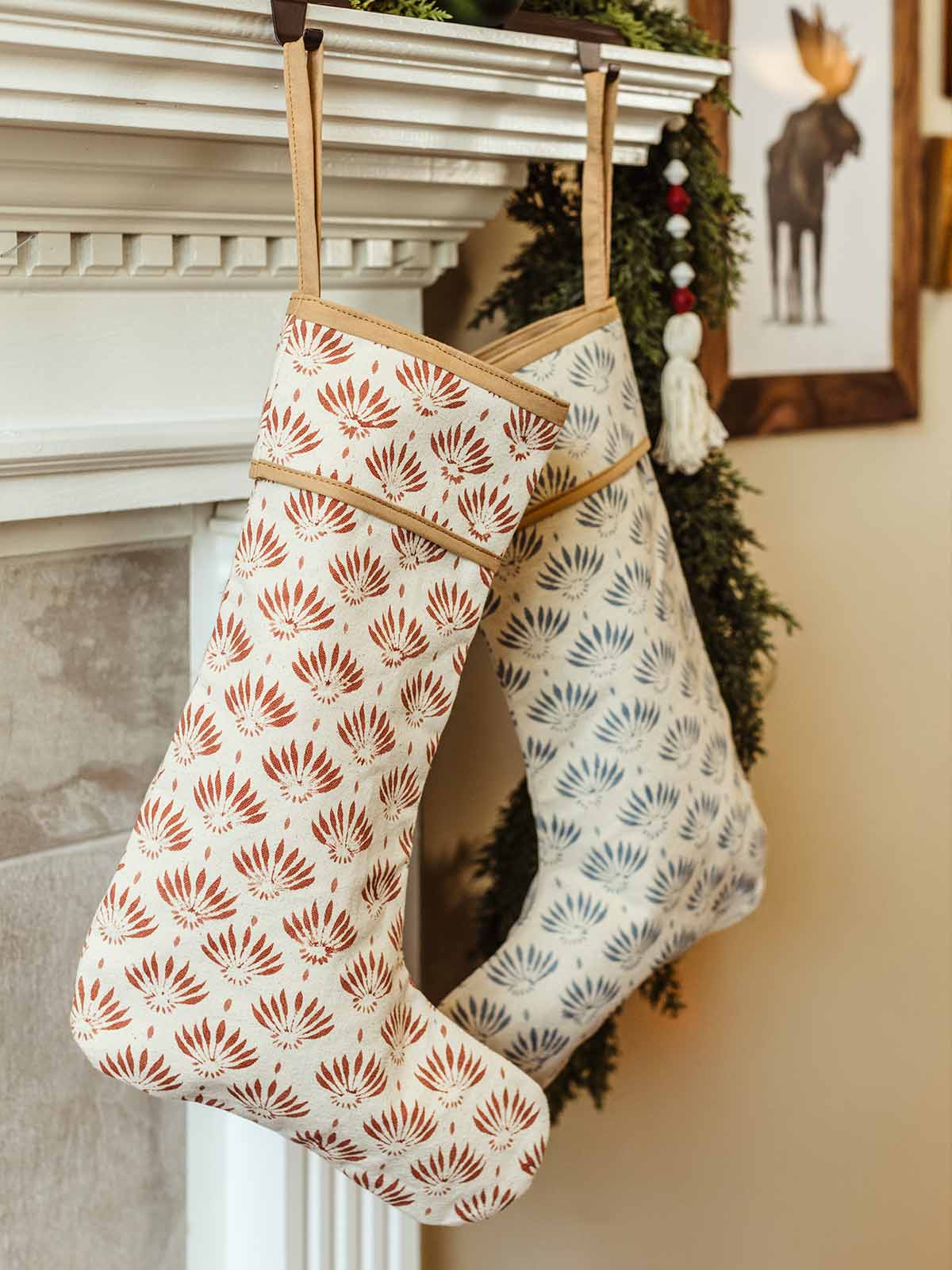 Close-up of two artisan-made Christmas stockings with botanical block print patterns, one in red and one in blue, hanging from a decorated mantel with evergreen garland and ornaments.