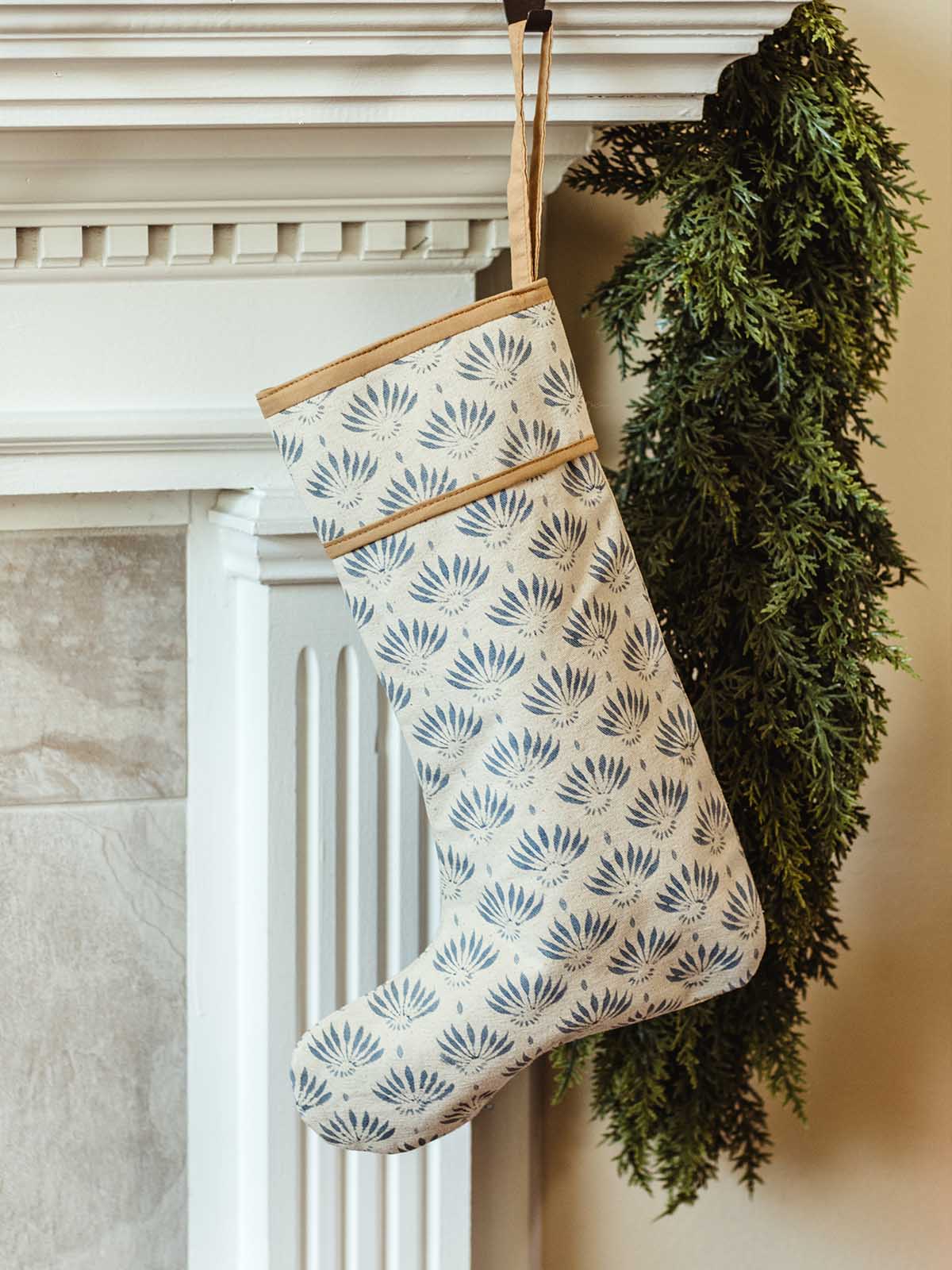Single blue hand-block printed Christmas stocking hanging on a white fireplace mantel with lush green garland, perfect for sustainable holiday décor.