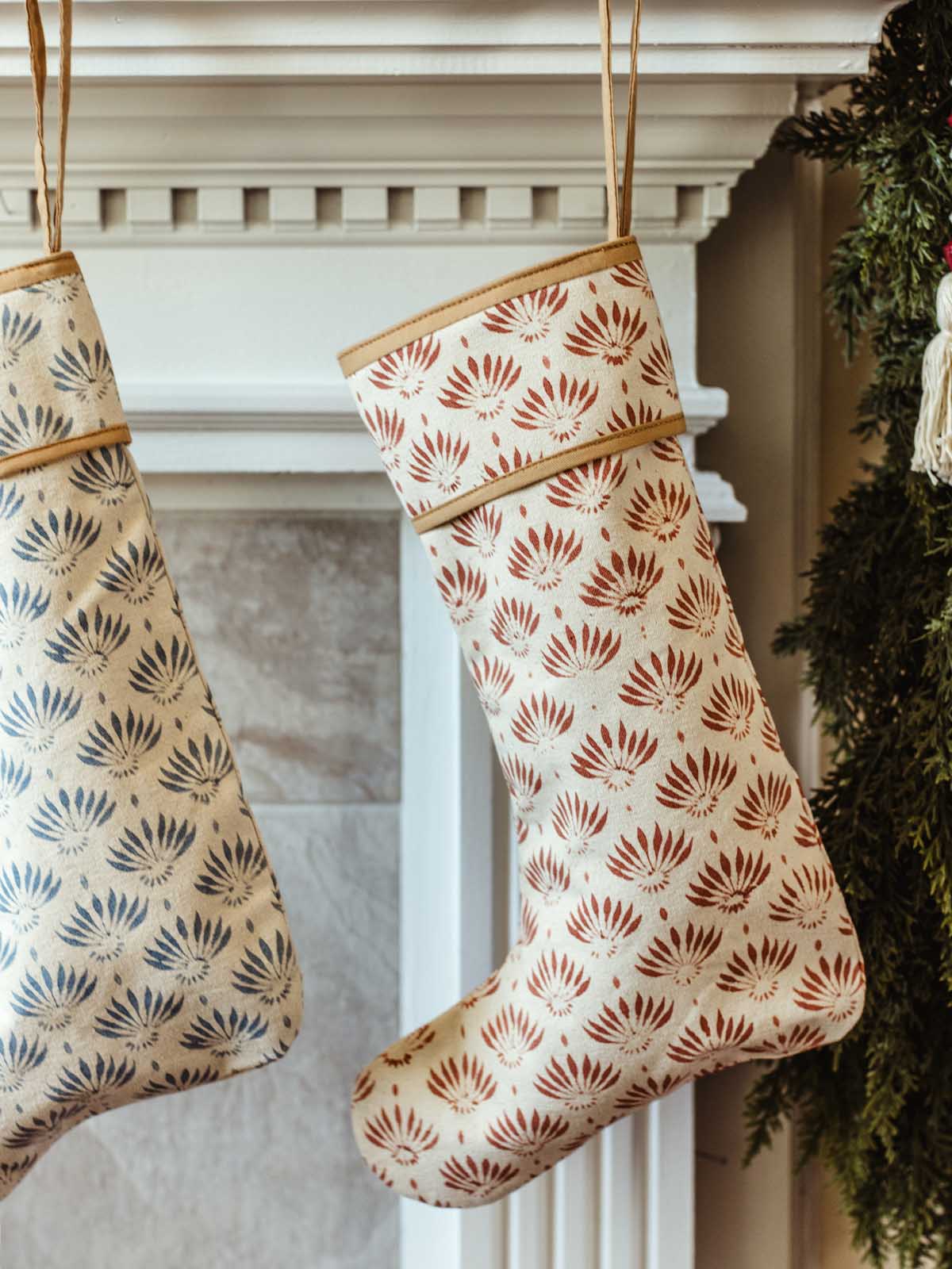 Red and blue hand-block printed Christmas stockings hanging side by side on a white mantel adorned with evergreen garland, wooden bead garland, and festive ornaments.