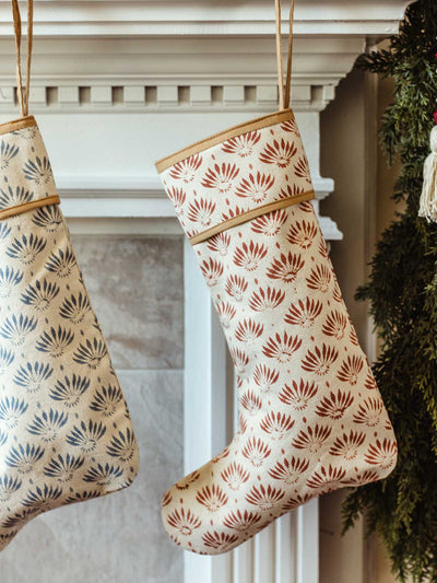 Red and blue hand-block printed Christmas stockings hanging side by side on a white mantel adorned with evergreen garland, wooden bead garland, and festive ornaments.