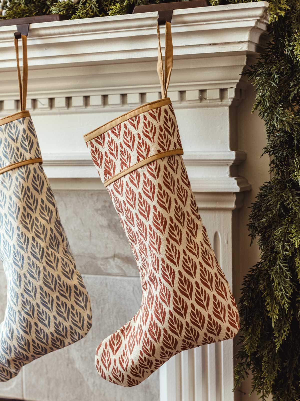 Red hand-block printed Christmas stocking with modern leaf pattern hanging from a white fireplace mantel, paired with a blue leaf-print stocking, styled with evergreen garland for cozy holiday décor.