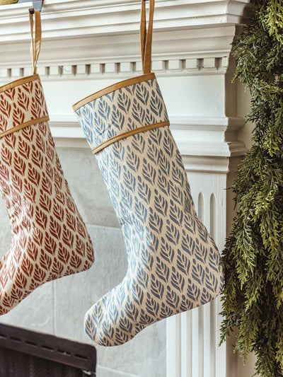 Blue hand-block printed Christmas stocking with leaf motif hanging beside a red stocking on a festive white mantel with garland and gold ornaments.