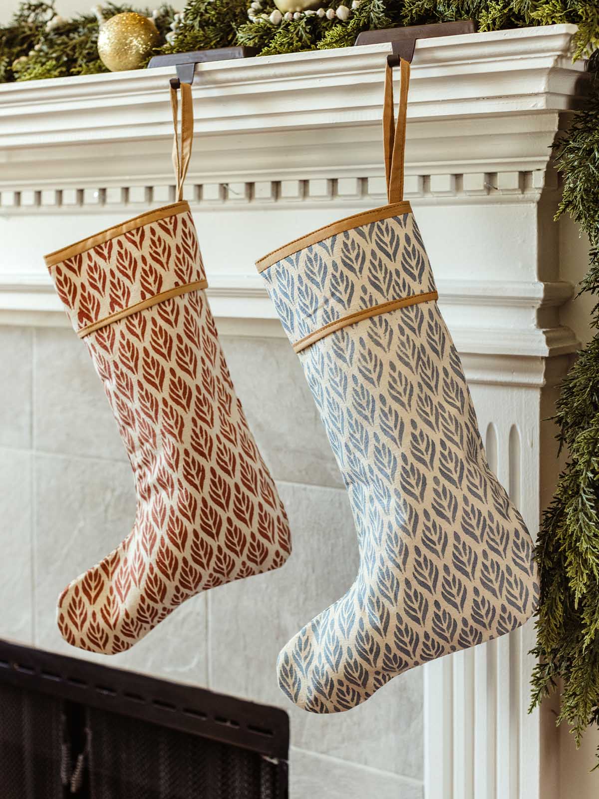 Pair of artisan-made Christmas stockings with red and blue leaf block prints hanging on a white fireplace mantel decorated with greenery, wood bead garland, and ornaments.